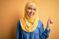 Young beautiful woman with curly hair wearing arab traditional hijab over yellow background with a big smile on face, pointing