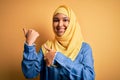 Young beautiful woman with curly hair wearing arab traditional hijab over yellow background Pointing to the back behind with hand
