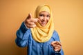 Young beautiful woman with curly hair wearing arab traditional hijab over yellow background pointing to you and the camera with