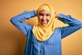 Young beautiful woman with curly hair wearing arab traditional hijab over yellow background relaxing and stretching, arms and