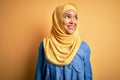 Young beautiful woman with curly hair wearing arab traditional hijab over yellow background smiling looking to the side and