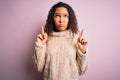 Young beautiful woman with curly hair wearing casual sweater standing over pink background Pointing up looking sad and upset, Royalty Free Stock Photo