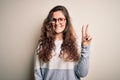 Young beautiful woman with curly hair wearing sweater and glasses over white background smiling with happy face winking at the Royalty Free Stock Photo