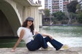 young, beautiful woman with dark, curly hair and an upturned nose is wearing a cap and sitting by a river. The woman is posing for