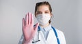 A young beautiful woman doctor in a white coat and pink sterile gloves shows a stop gesture on gray background. Royalty Free Stock Photo