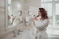 Young beautiful woman with dyed hair in white bathrobe in bathroom dries hair with hairdryer and smiles. Royalty Free Stock Photo