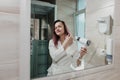 Young beautiful woman with dyed hair in white bathrobe in bathroom dries hair with hairdryer and smiles. Royalty Free Stock Photo