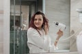 Young beautiful woman with dyed hair in white bathrobe in bathroom dries hair with hairdryer and smiles. Royalty Free Stock Photo