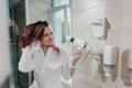 Young beautiful woman with dyed hair in white bathrobe in bathroom dries hair with hairdryer and smiles. Royalty Free Stock Photo