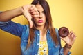 Young beautiful woman eating chocolate donut over yellow background with angry face, negative sign showing dislike with thumbs