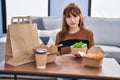 Young beautiful woman eating delivery food at the living room thinking attitude and sober expression looking self confident