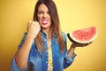 Young beautiful woman eating fresh healthy watermelon slice over yellow background annoyed and frustrated shouting with anger, Royalty Free Stock Photo