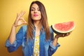 Young beautiful woman eating fresh healthy watermelon slice over yellow background doing ok sign with fingers, excellent symbol Royalty Free Stock Photo