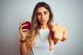 Young beautiful woman eating red apple over grey  background pointing with finger to the camera and to you, hand sign, Royalty Free Stock Photo