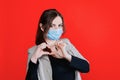 Young beautiful woman in face medical mask shows heart jesture, bright red background.