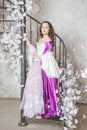 Beautiful woman in fantasy white and purple rococo style medieval dress standing on the stairway with white flowers