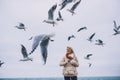 Young beautiful woman feeds seagulls on the sea. Pretty female wearing coat, scarf and watching flying seagulls by the sea in the Royalty Free Stock Photo