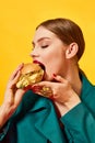 Young beautiful woman in green coat eating, biting cheeseburger with necklaces over yellow background. Luxury food. Food