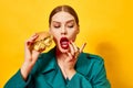 Young beautiful woman in green coat eating cheeseburger with necklaces over yellow background. Luxury food. Food pop art