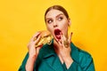 Young beautiful woman in green coat with red lipstick eating cheeseburger with necklaces over yellow background. Food