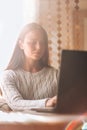 Young beautiful woman in a grey cozy sweater works for a computer from a home with a laptop on a wooden desk as a Royalty Free Stock Photo