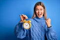 Young beautiful woman holding alarm clock standing over isolated blue background screaming proud and celebrating victory and Royalty Free Stock Photo