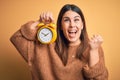 Young beautiful woman holding alarm clock standing over isolated orange background screaming proud and celebrating victory and Royalty Free Stock Photo