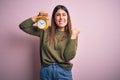 Young beautiful woman holding alarm clock standing over isolated pink background screaming proud and celebrating victory and Royalty Free Stock Photo