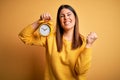 Young beautiful woman holding alarm clock standing over isolated yellow background screaming proud and celebrating victory and Royalty Free Stock Photo