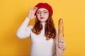 Young beautiful woman holding bag of fresh baguette over yellow background, poses with hand on head for mistake, remember error, Royalty Free Stock Photo