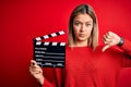 Young beautiful woman holding clapboard standing over isolated red background with angry face, negative sign showing dislike with