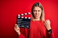 Young beautiful woman holding clapboard standing over isolated red background annoyed and frustrated shouting with anger, crazy Royalty Free Stock Photo