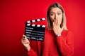 Young beautiful woman holding clapboard standing over isolated red background cover mouth with hand shocked with shame for Royalty Free Stock Photo