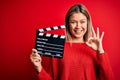 Young beautiful woman holding clapboard standing over isolated red background doing ok sign with fingers, excellent symbol Royalty Free Stock Photo