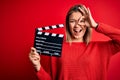 Young beautiful woman holding clapboard standing over isolated red background with happy face smiling doing ok sign with hand on Royalty Free Stock Photo