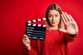 Young beautiful woman holding clapboard standing over isolated red background with open hand doing stop sign with serious and Royalty Free Stock Photo