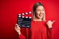 Young beautiful woman holding clapboard standing over isolated red background pointing and showing with thumb up to the side with