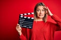 Young beautiful woman holding clapboard standing over isolated red background stressed with hand on head, shocked with shame and Royalty Free Stock Photo