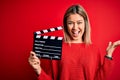 Young beautiful woman holding clapboard standing over isolated red background very happy and excited, winner expression Royalty Free Stock Photo