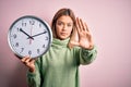 Young beautiful woman holding clock standing over isolated pink background with open hand doing stop sign with serious and Royalty Free Stock Photo