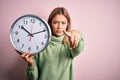 Young beautiful woman holding clock standing over isolated pink background pointing with finger to the camera and to you, hand Royalty Free Stock Photo