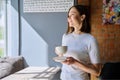 Young beautiful woman holding cup of coffee, looking out window, gray wall copy space Royalty Free Stock Photo