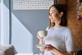 Young beautiful woman holding cup of coffee, looking out window, gray wall copy space Royalty Free Stock Photo