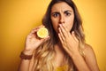 Young beautiful woman holding half lemon over yellow isolated background cover mouth with hand shocked with shame for mistake,