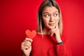 Young beautiful woman holding heart card standing over isolated red background serious face thinking about question, very confused Royalty Free Stock Photo