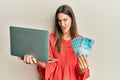 Young beautiful woman holding laptop and holding brazilian real in shock face, looking skeptical and sarcastic, surprised with