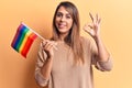 Young beautiful woman holding lgtbq flag doing ok sign with fingers, smiling friendly gesturing excellent symbol