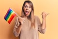 Young beautiful woman holding lgtbq flag pointing thumb up to the side smiling happy with open mouth