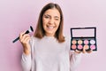 Young beautiful woman holding makeup brush and blush winking looking at the camera with sexy expression, cheerful and happy face Royalty Free Stock Photo