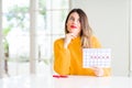 Young beautiful woman holding menstruation calendar at home serious face thinking about question, very confused idea